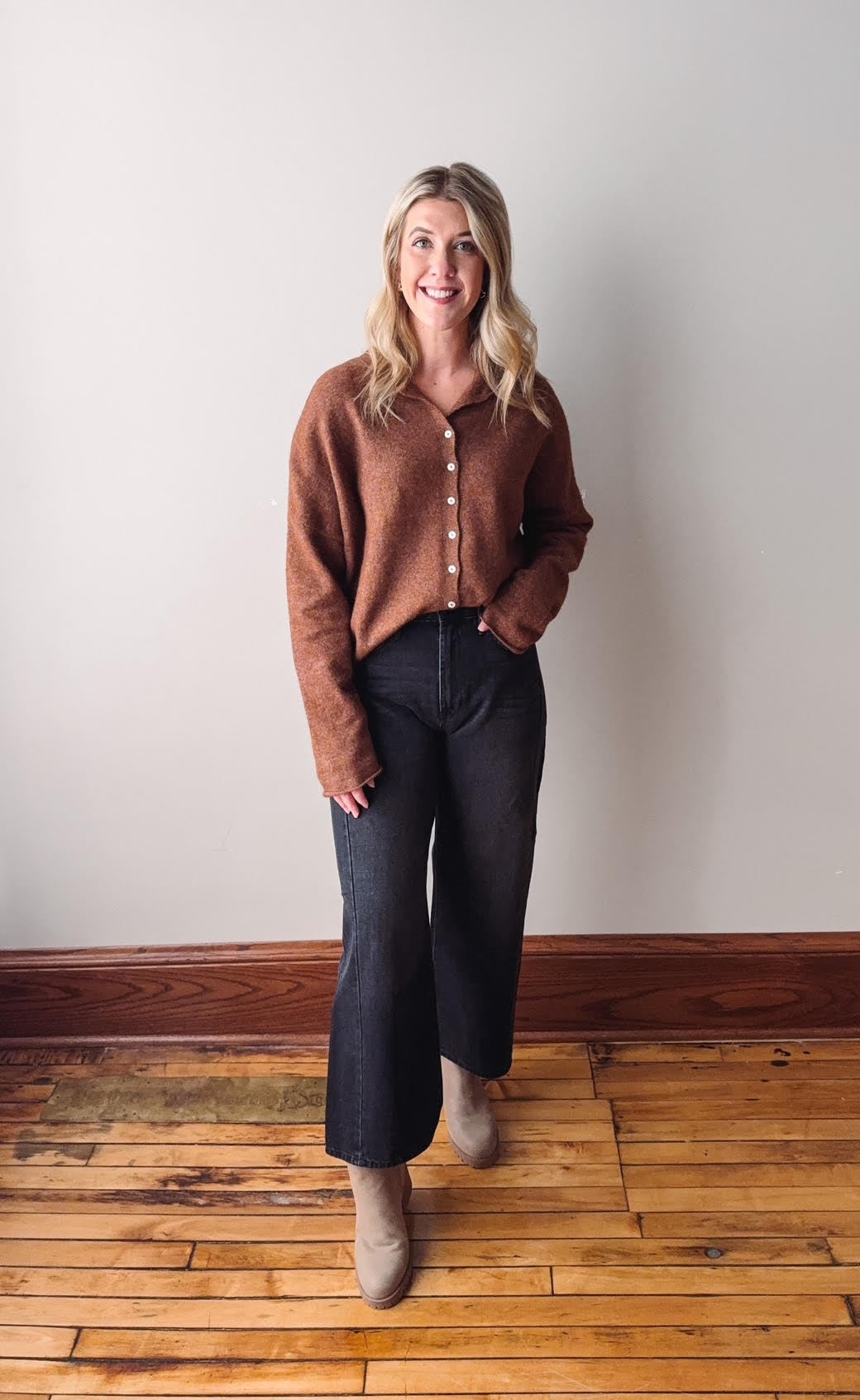 Woman wearing a brown sweater and dark jeans standing on a wooden floor with a white wall background