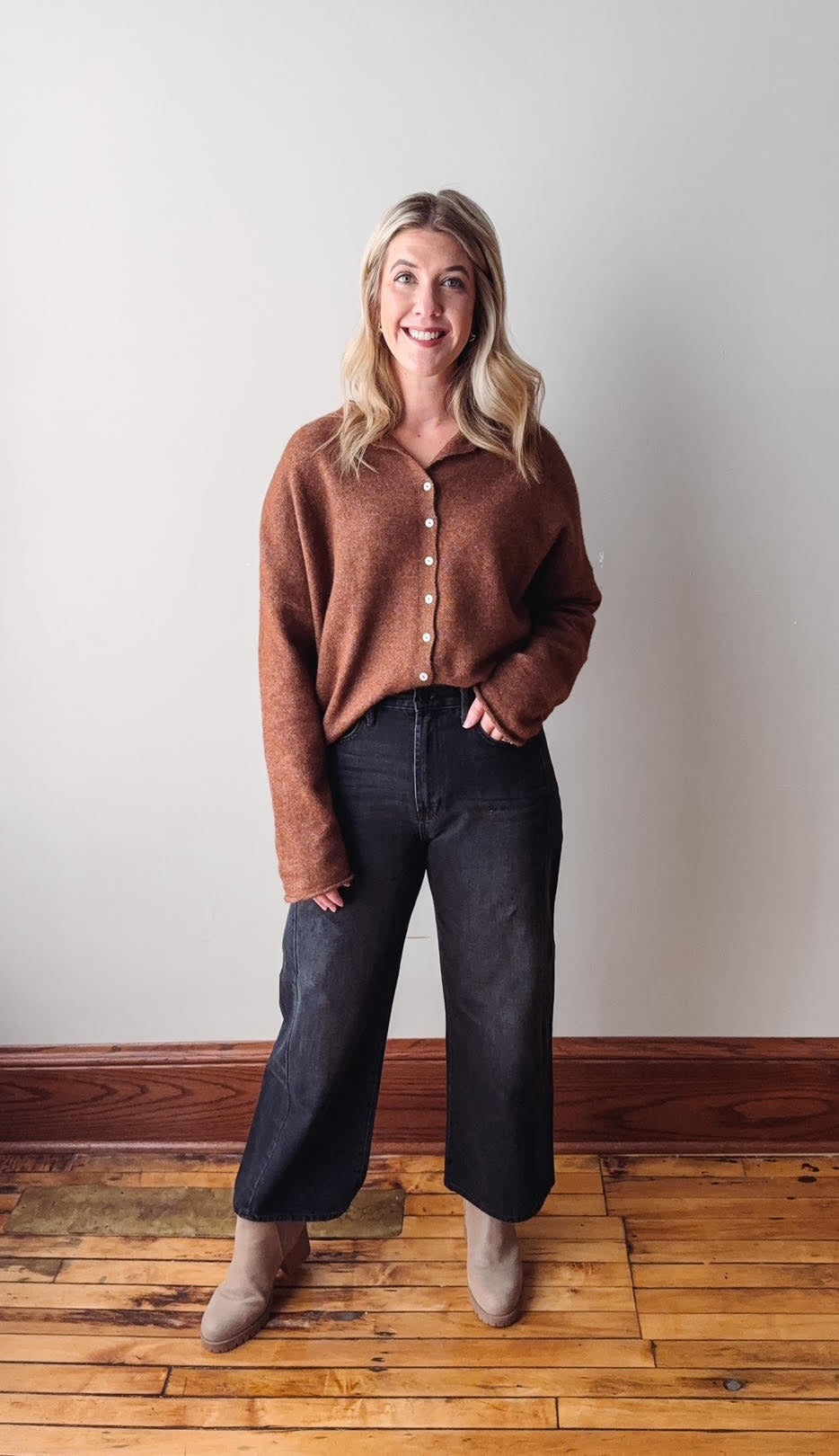 Woman wearing a brown sweater and dark jeans standing on a wooden floor against a white wall.