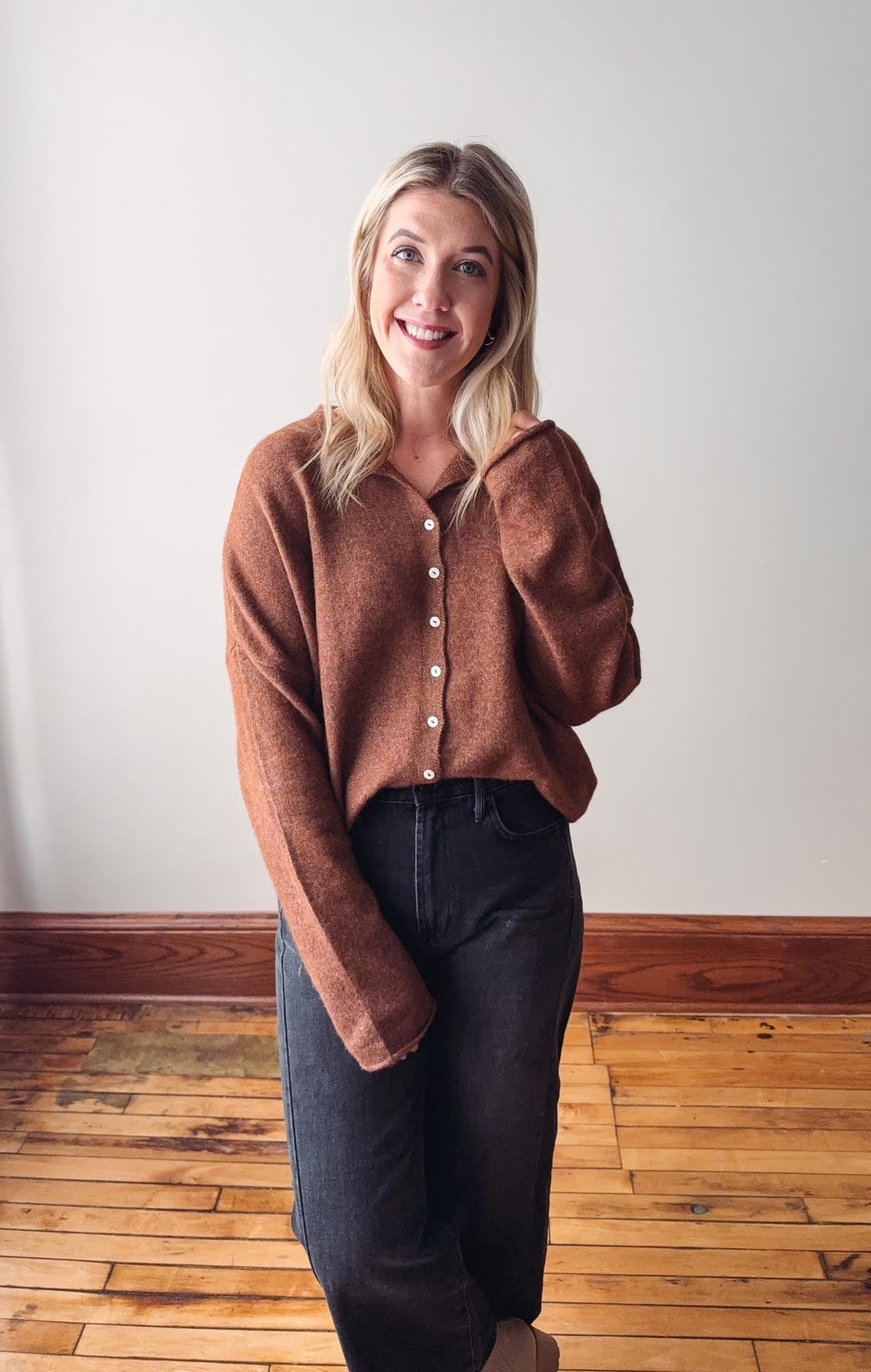 Woman wearing a brown sweater and dark jeans standing on a wooden floor with a white wall background