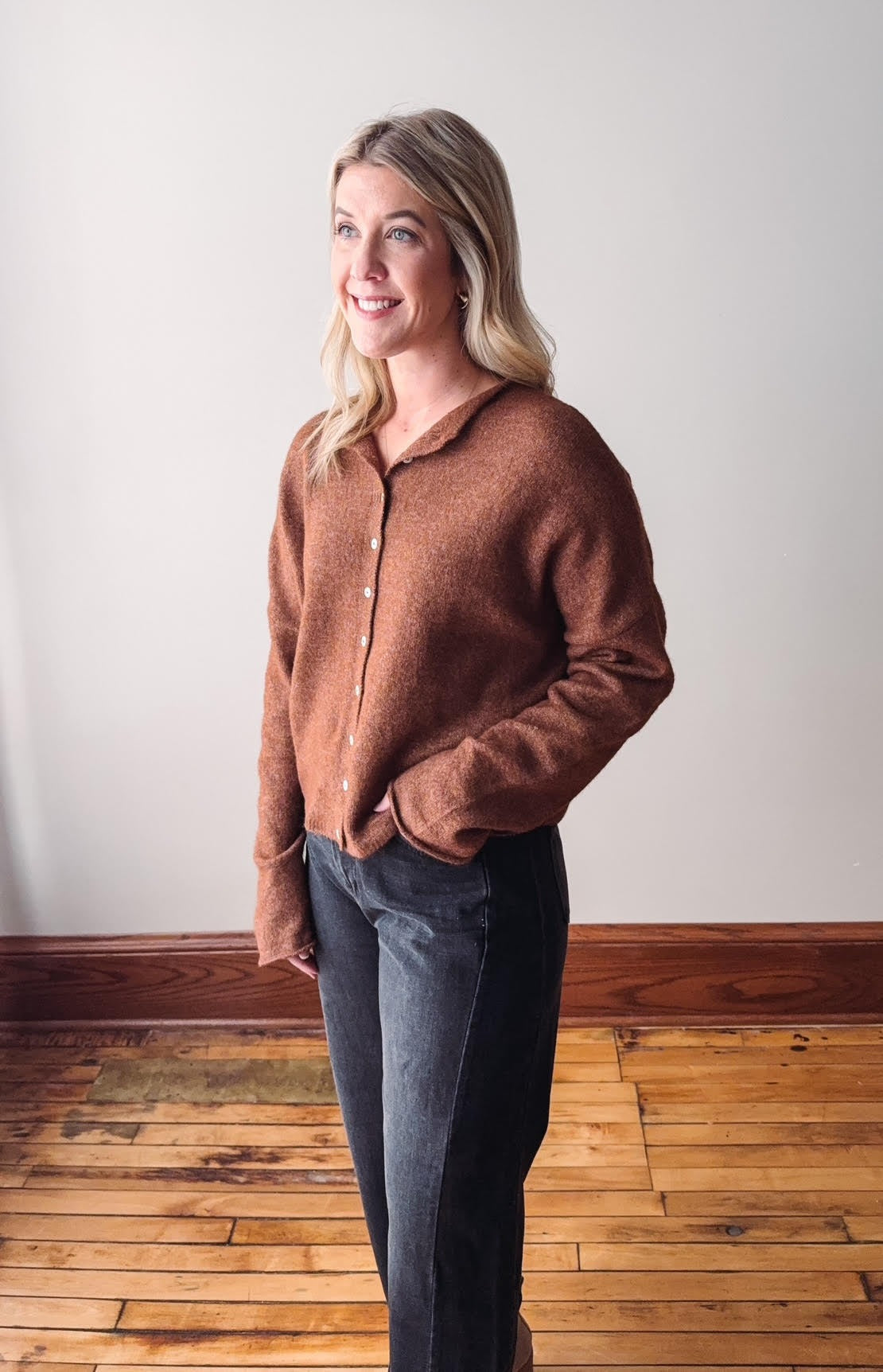 Woman wearing a brown sweater and dark jeans standing on a wooden floor with a white wall background