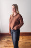 Woman wearing a brown sweater and dark jeans standing on a wooden floor with a white wall background