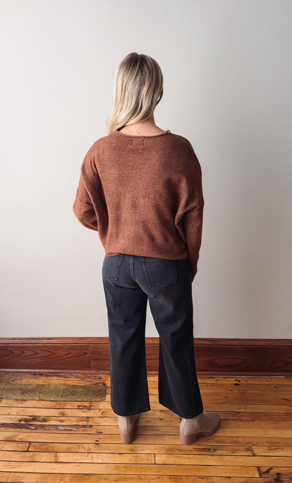 Person wearing a brown sweater and dark jeans standing on a wooden floor with a white wall background