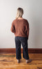 Person wearing a brown sweater and dark jeans standing on a wooden floor with a white wall background