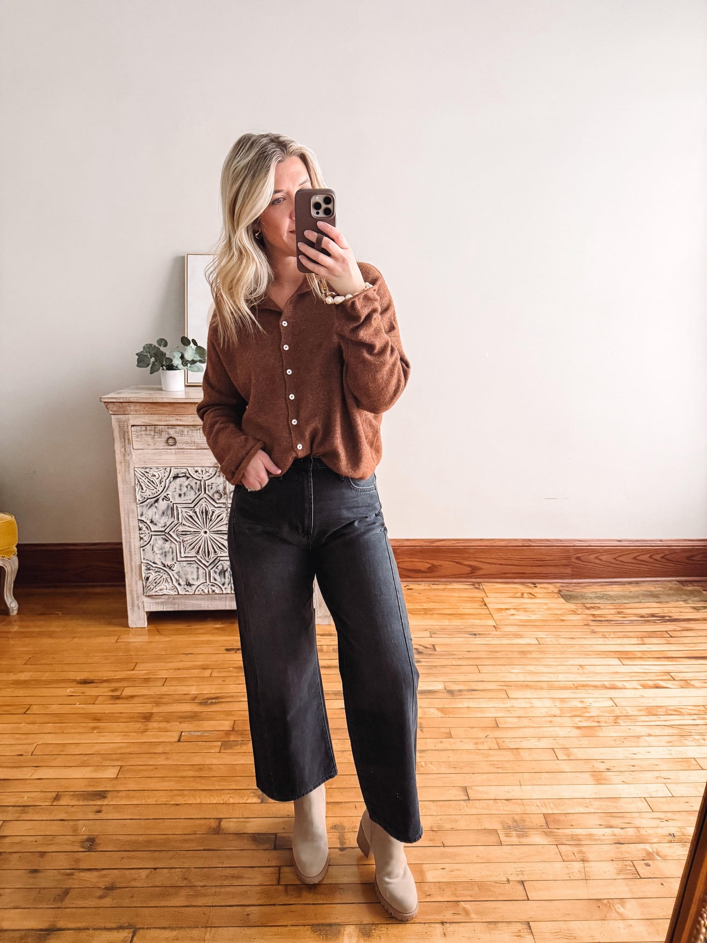Person taking a mirror selfie wearing a brown shirt and black jeans in a room with wooden flooring.