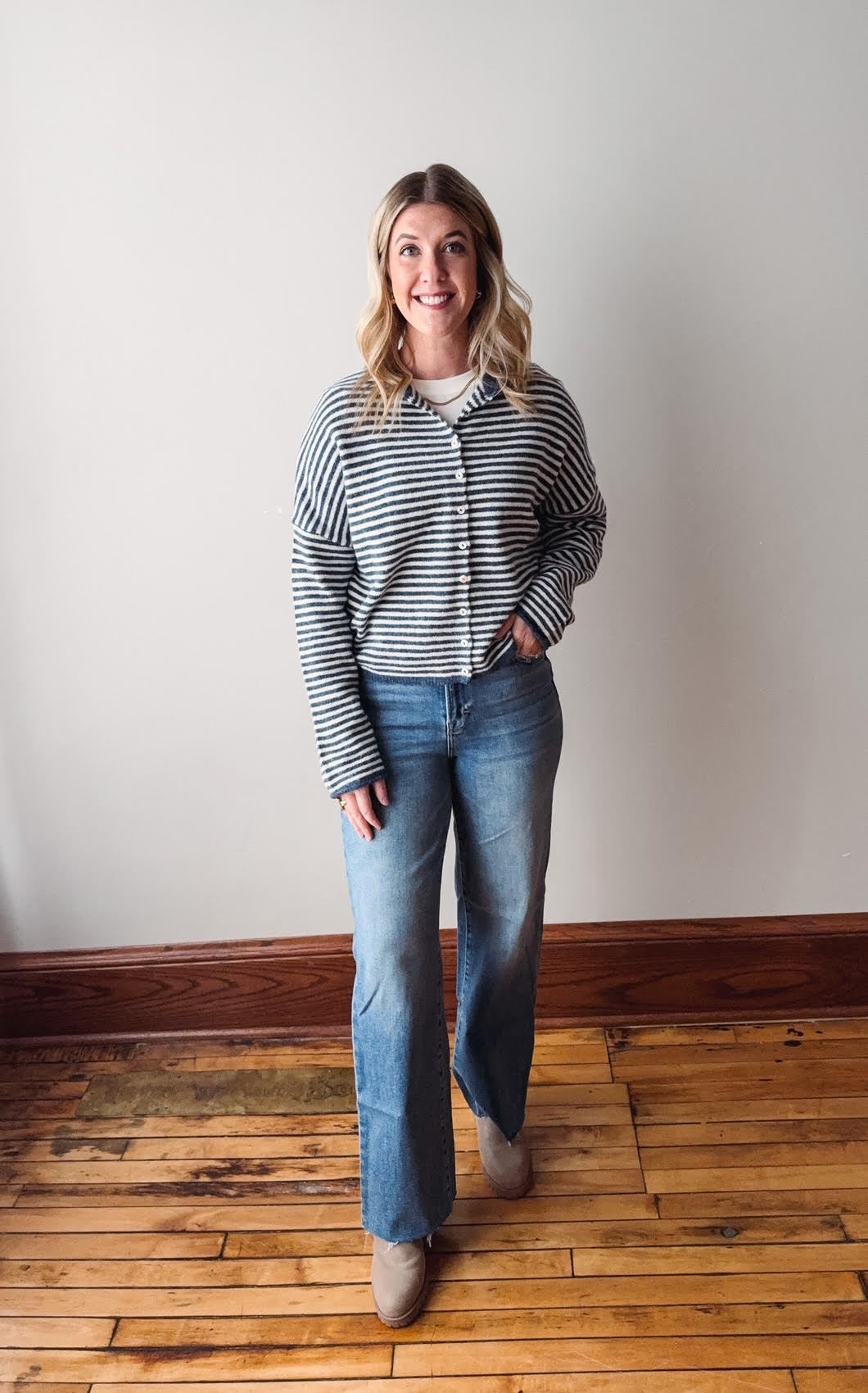 Woman wearing a striped shirt and jeans standing on a wooden floor with a white wall background