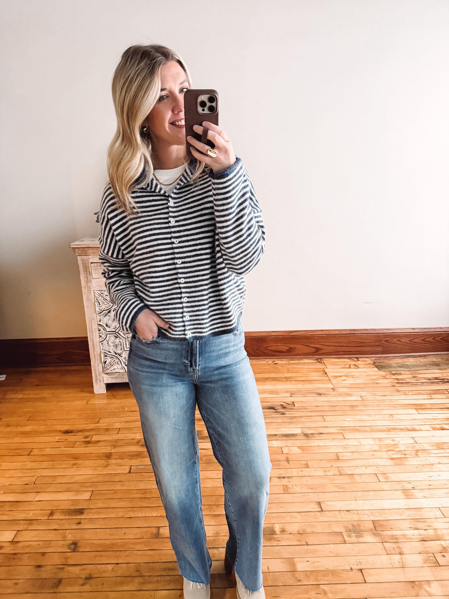 Woman taking a mirror selfie wearing a striped shirt and jeans in a room with wooden flooring.