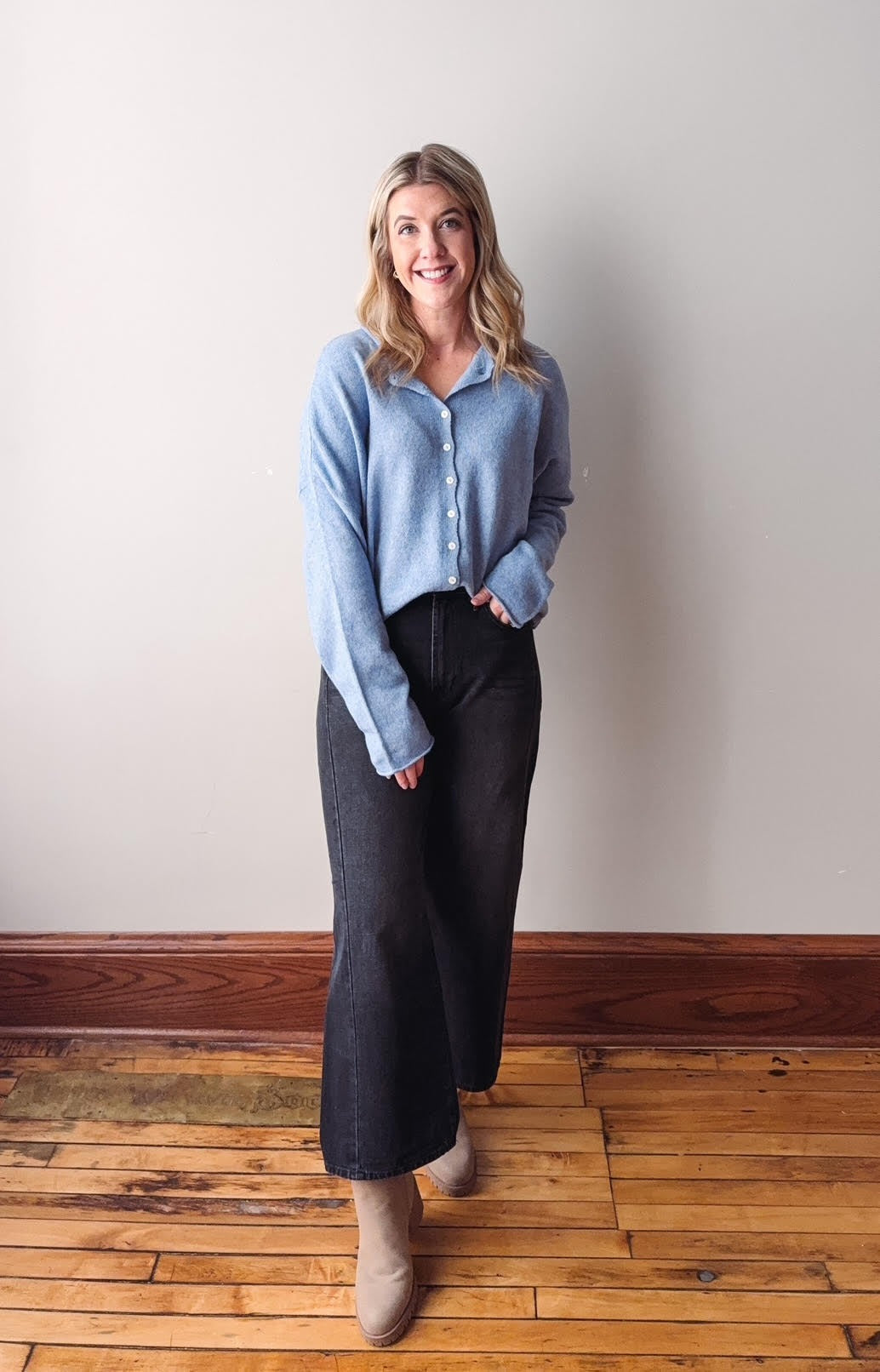 Woman wearing a light blue sweater and dark pants standing on a wooden floor against a white wall.