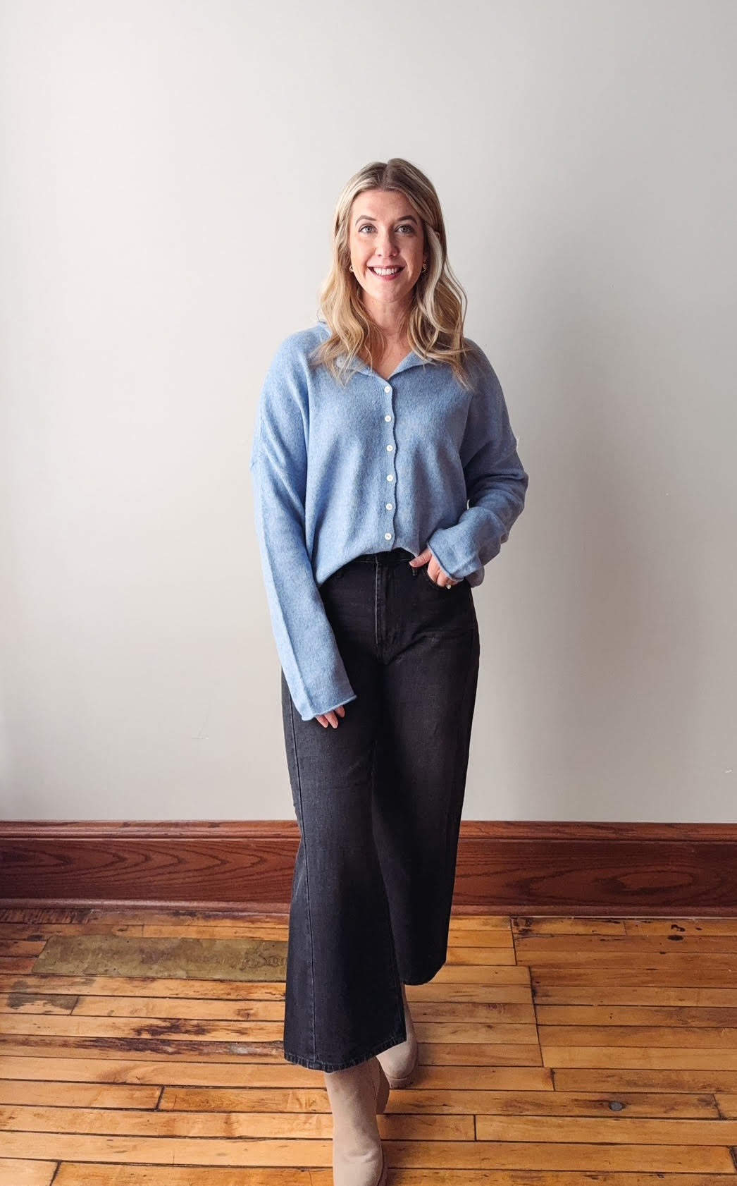 Woman wearing a light blue sweater and dark pants standing on a wooden floor against a white wall.