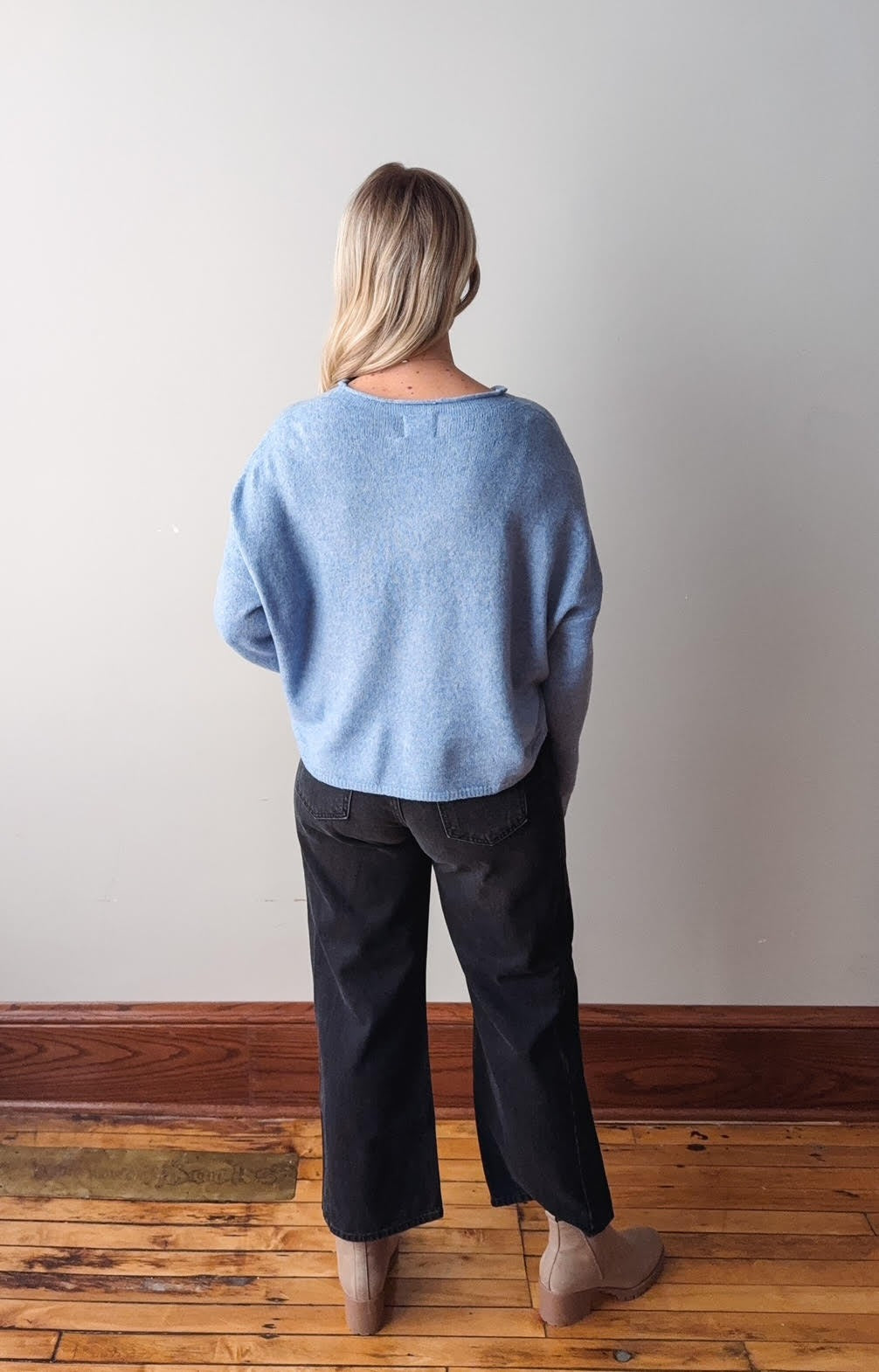 Person wearing a light blue sweater and dark pants standing against a white wall on a wooden floor.