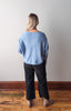 Person wearing a light blue sweater and dark pants standing against a white wall on a wooden floor.