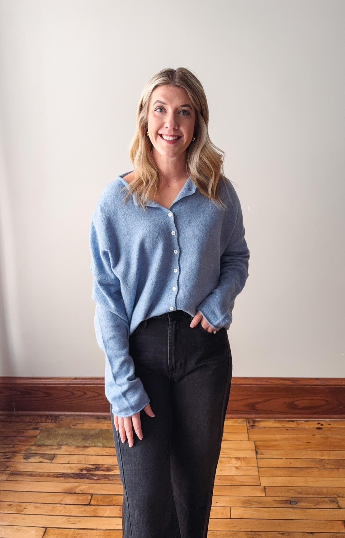 Woman wearing a light blue sweater and dark pants standing on a wooden floor with a white wall background