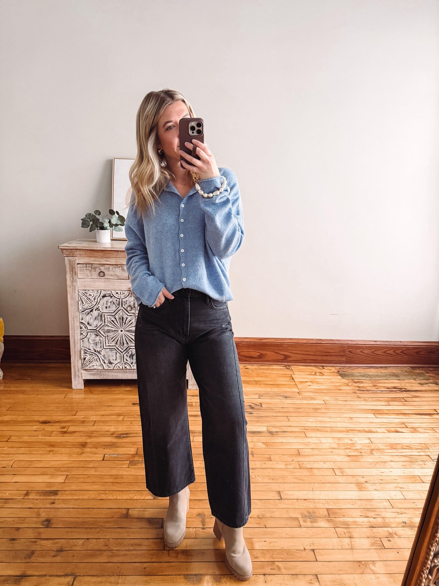 Person taking a mirror selfie wearing a blue shirt and black pants in a room with wooden flooring and a decorative cabinet.