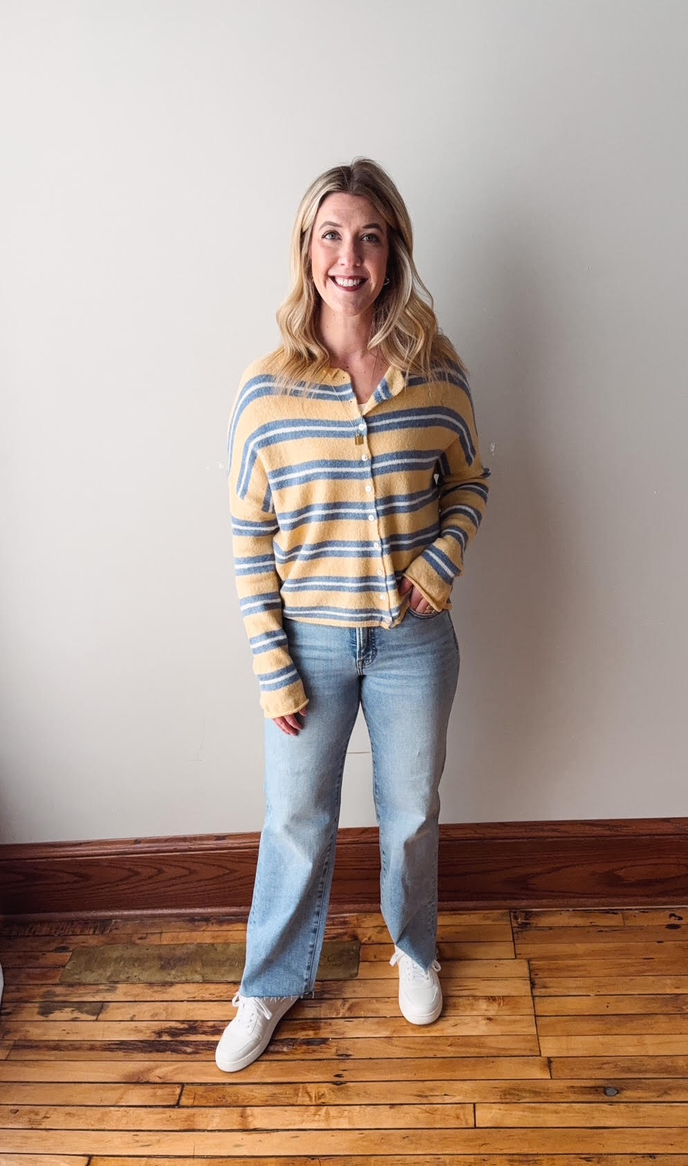 Woman wearing a striped shirt and jeans standing against a plain wall.
