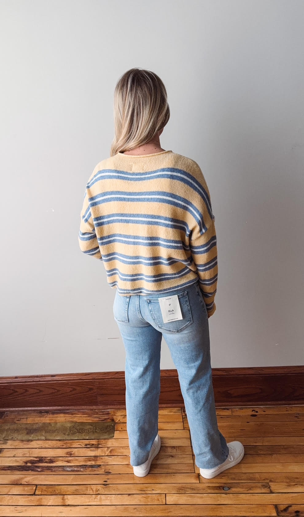 Person wearing a striped sweater and jeans standing on a wooden floor with a white wall background