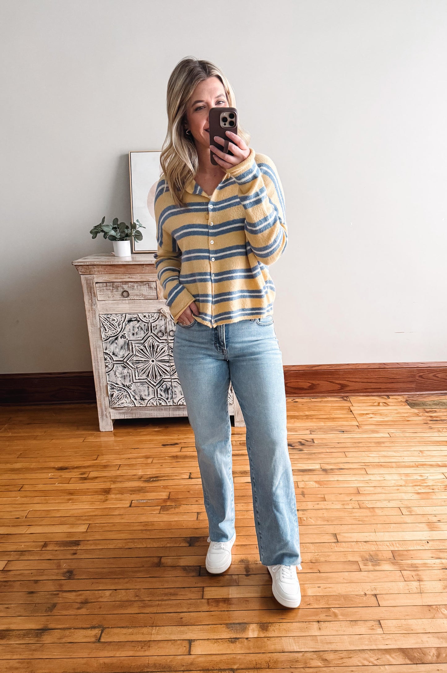 Woman taking a mirror selfie wearing a striped sweater and jeans in a room with wooden flooring and a decorative cabinet.