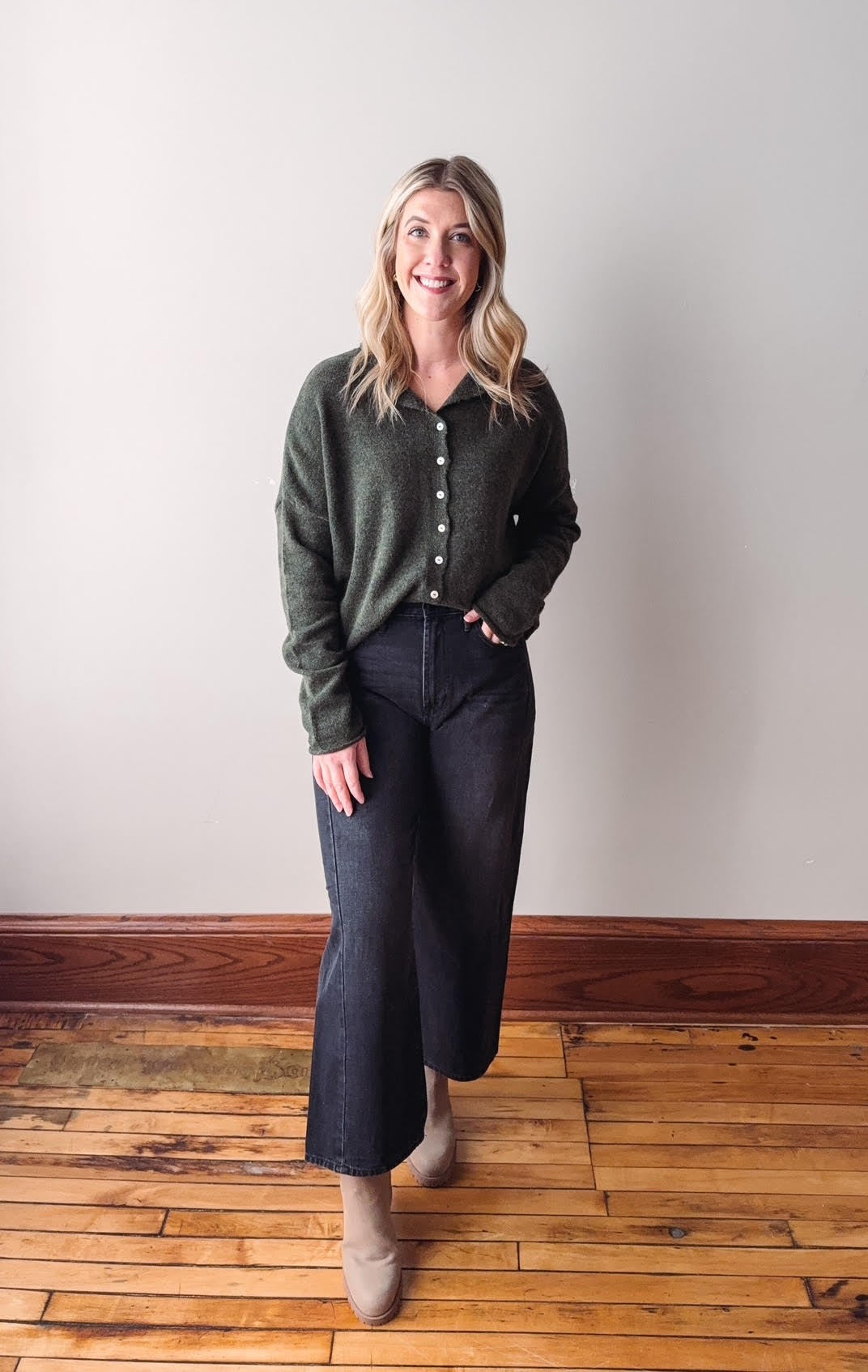 Woman wearing a green sweater and dark jeans standing on a wooden floor with a white wall background