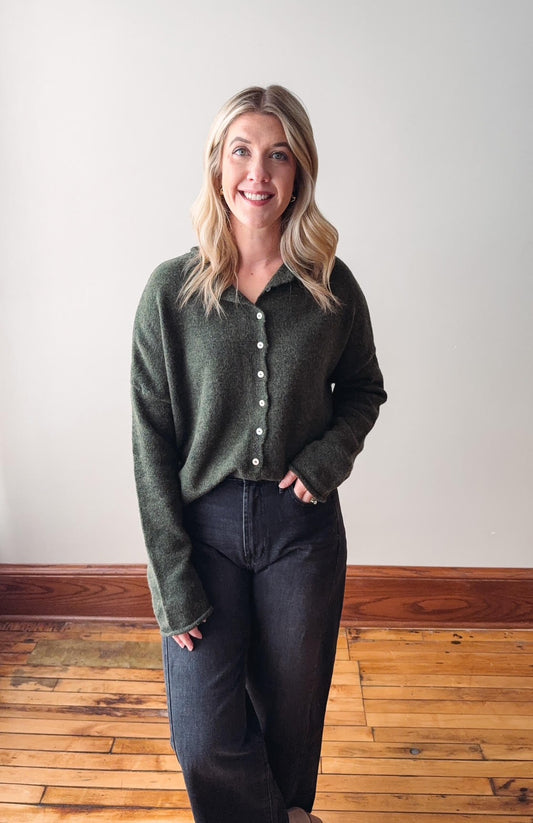 Woman wearing a green cardigan and dark pants standing on a wooden floor with a white wall background