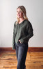 Woman wearing a green sweater and dark jeans standing on a wooden floor with a white wall background