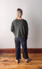 Person wearing a green sweater and dark pants standing on a wooden floor with a white wall background