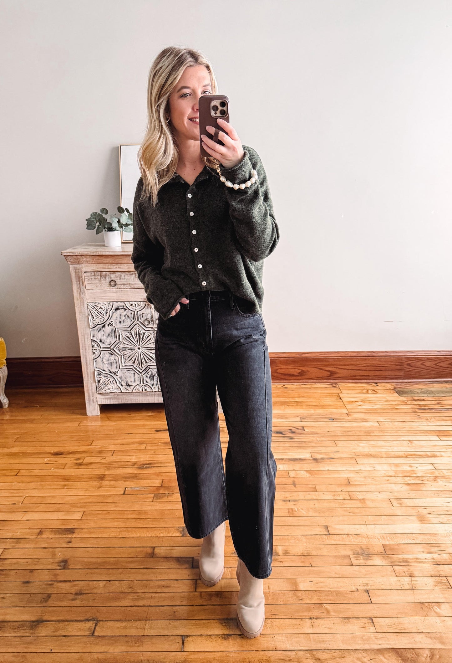 Woman taking a mirror selfie wearing a dark green shirt and black jeans in a room with wooden flooring.