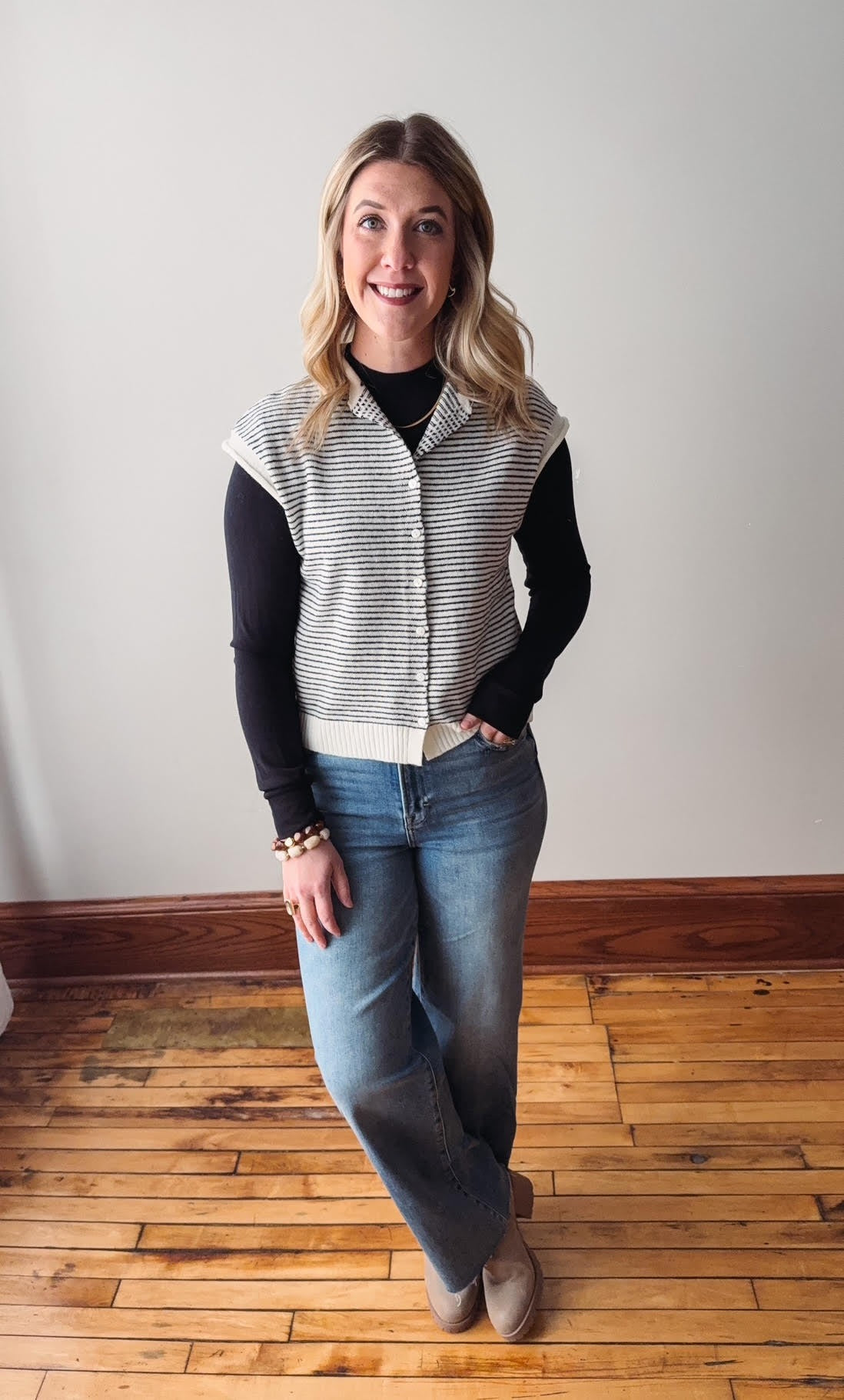 Woman wearing a black and white striped vest over a black shirt and blue jeans on a wooden floor.