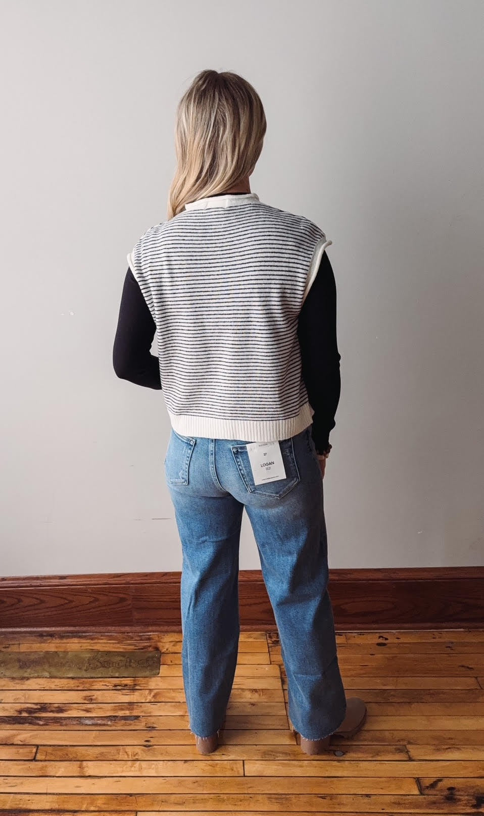 Person wearing a striped vest and blue jeans standing on a wooden floor against a plain wall.
