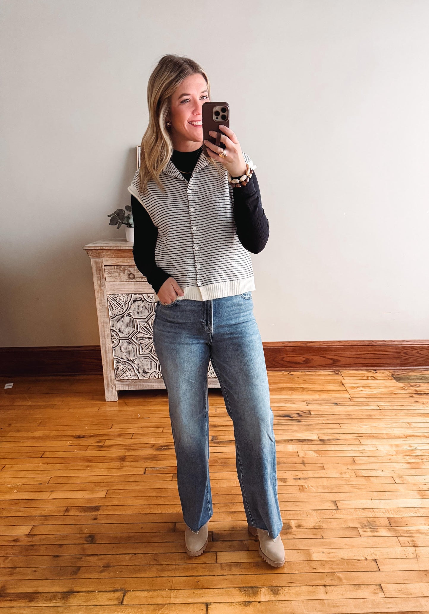 Woman taking a mirror selfie wearing a striped vest, black top, and blue jeans in a room with wooden flooring.