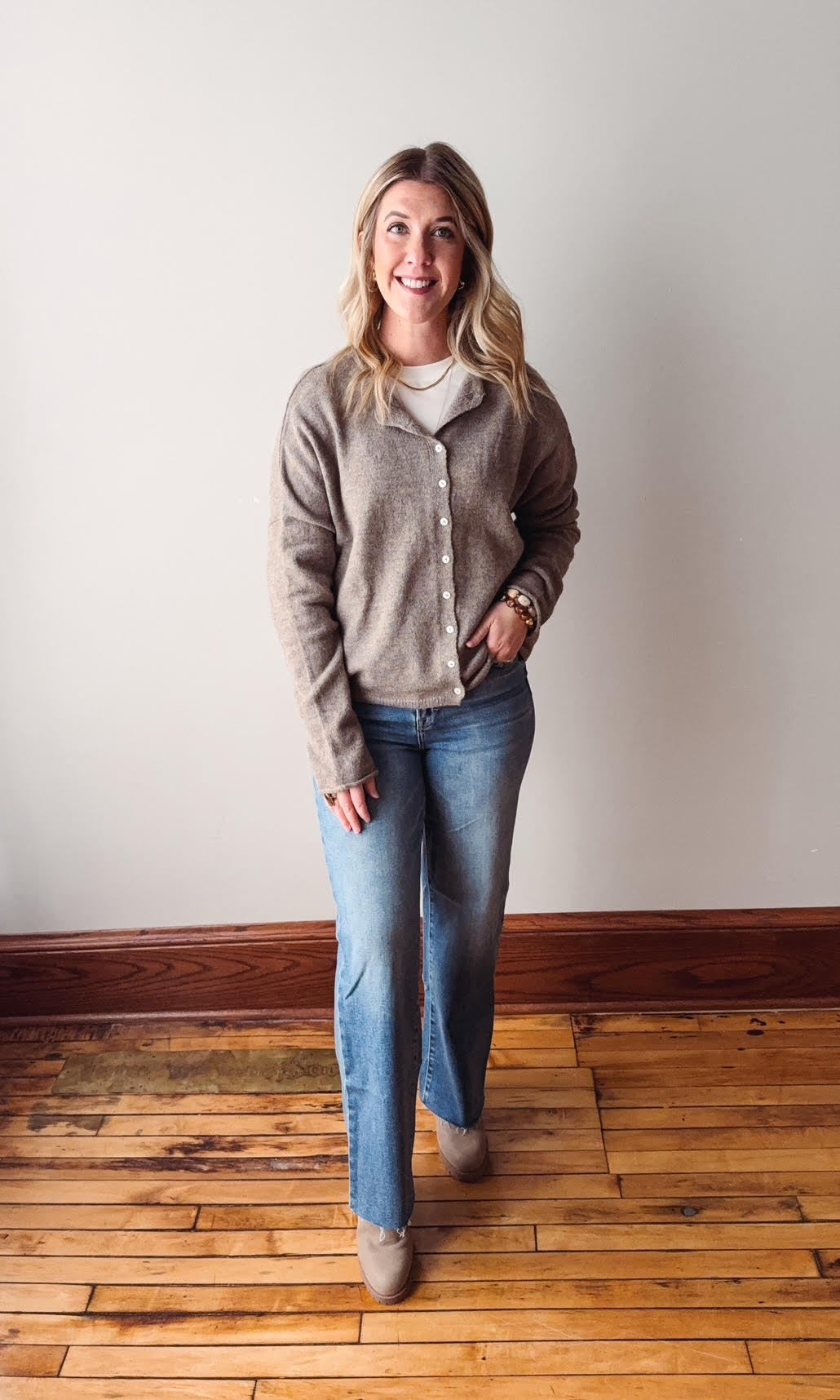 Woman wearing a gray cardigan and blue jeans standing on a wooden floor with a white wall background