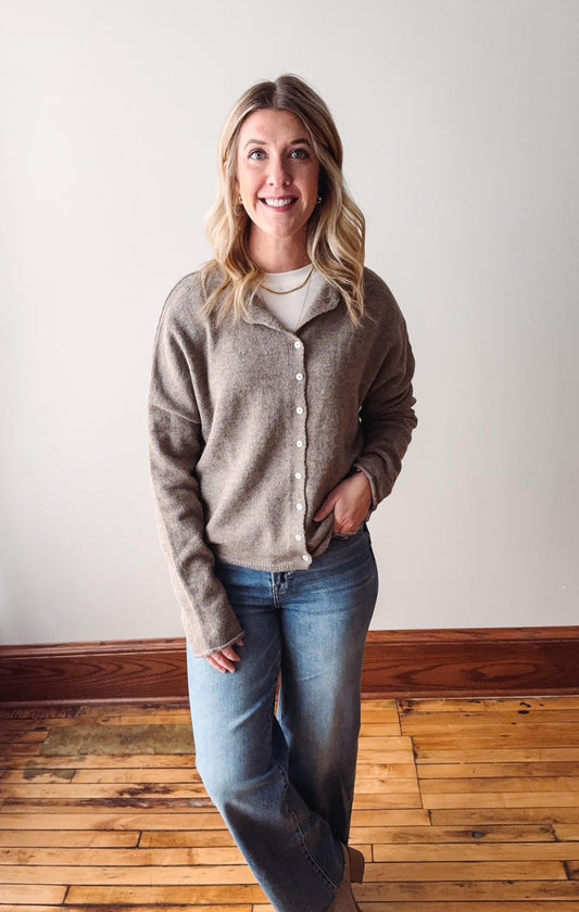 Woman wearing a gray cardigan and blue jeans standing on a wooden floor with a white wall background.