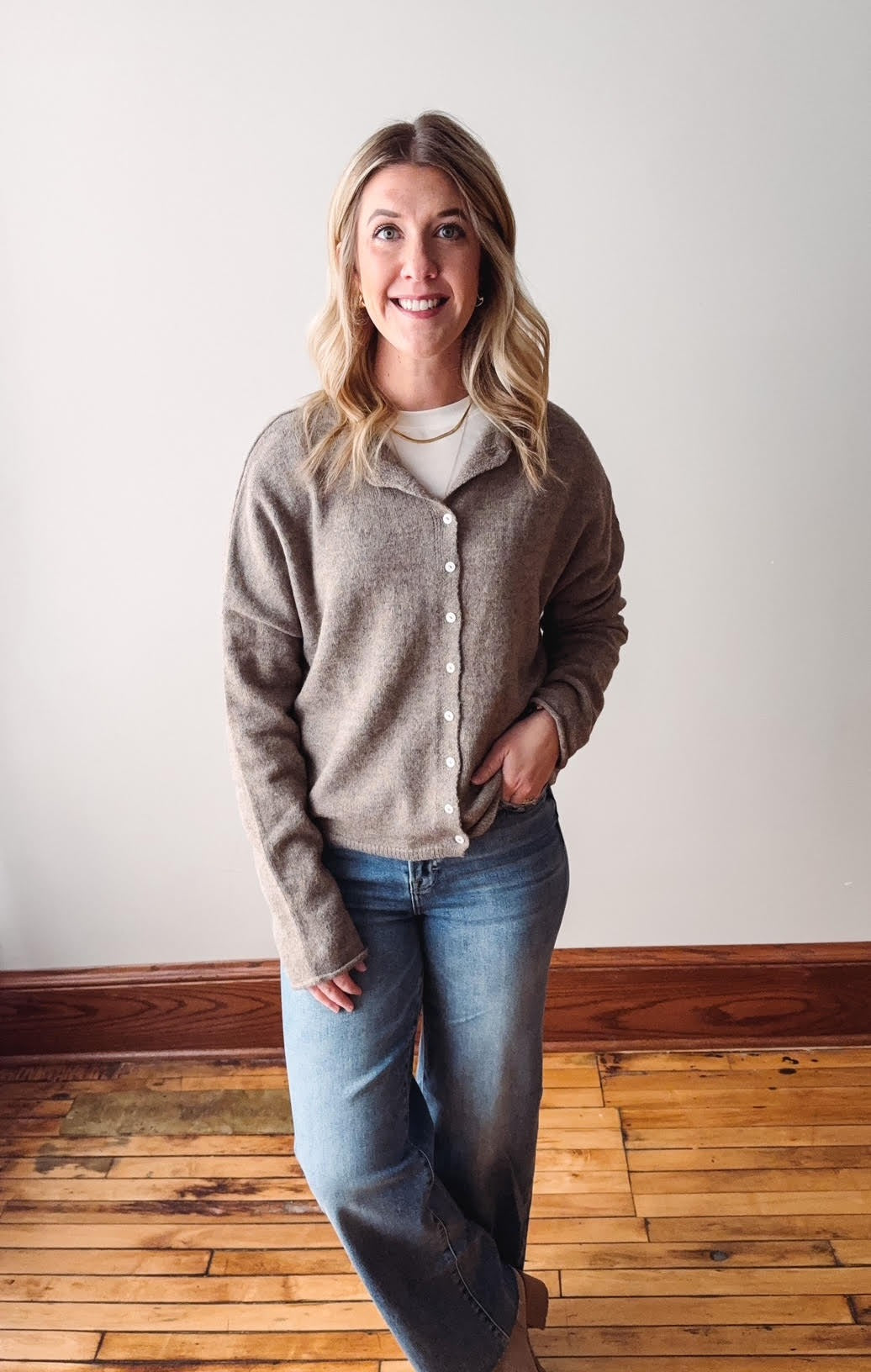 Woman wearing a gray cardigan and blue jeans standing on a wooden floor with a white wall background.