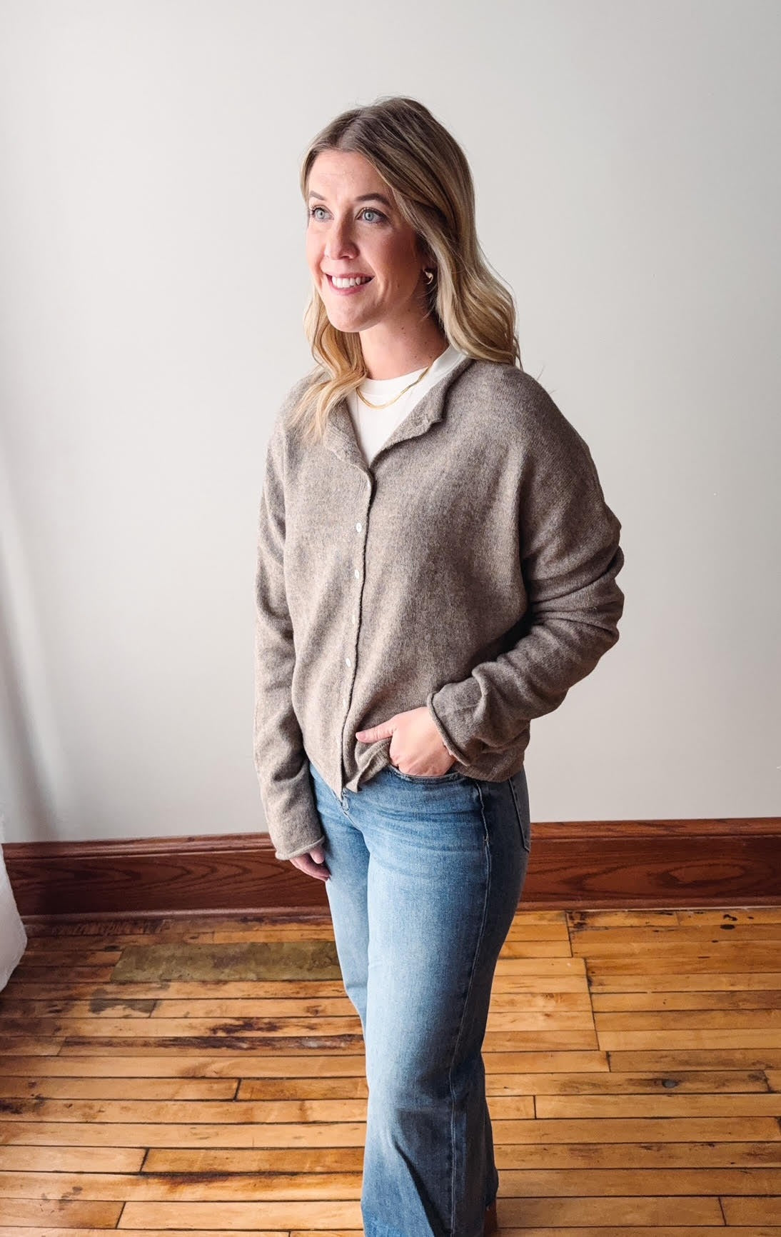 Woman wearing a brown jacket and blue jeans standing on a wooden floor.