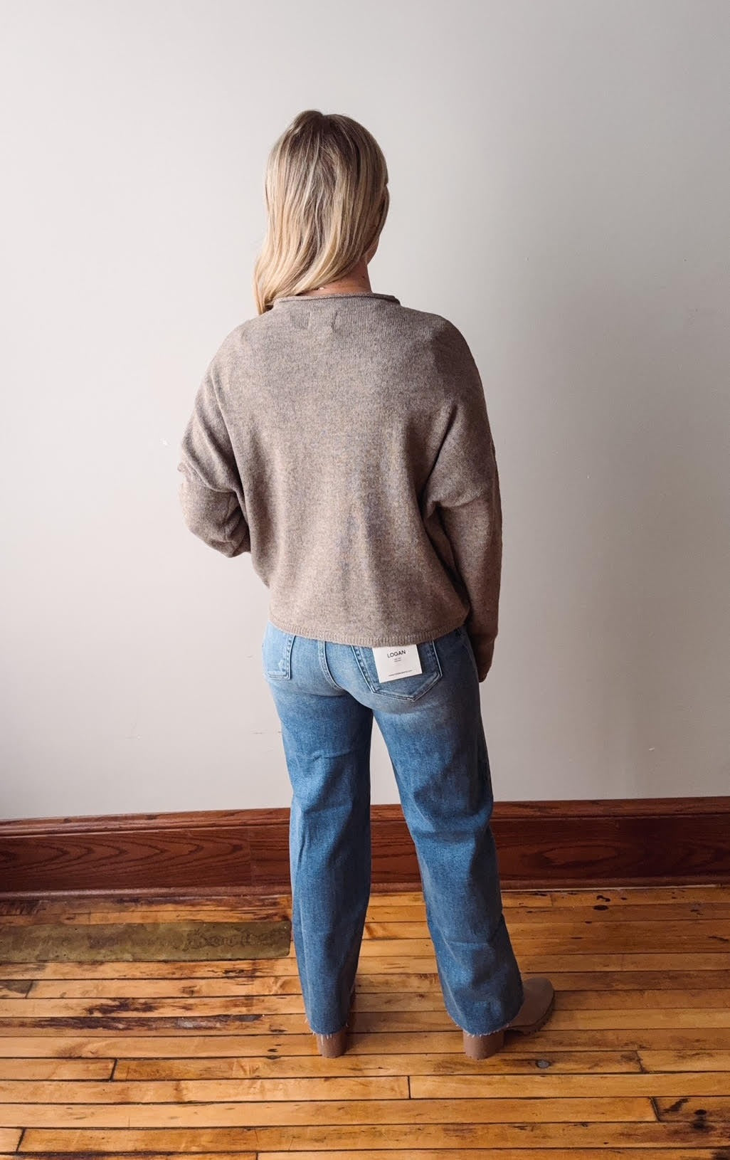 Person wearing a gray sweater and blue jeans standing on a wooden floor against a white wall.