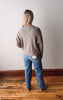 Person wearing a gray sweater and blue jeans standing on a wooden floor against a white wall.