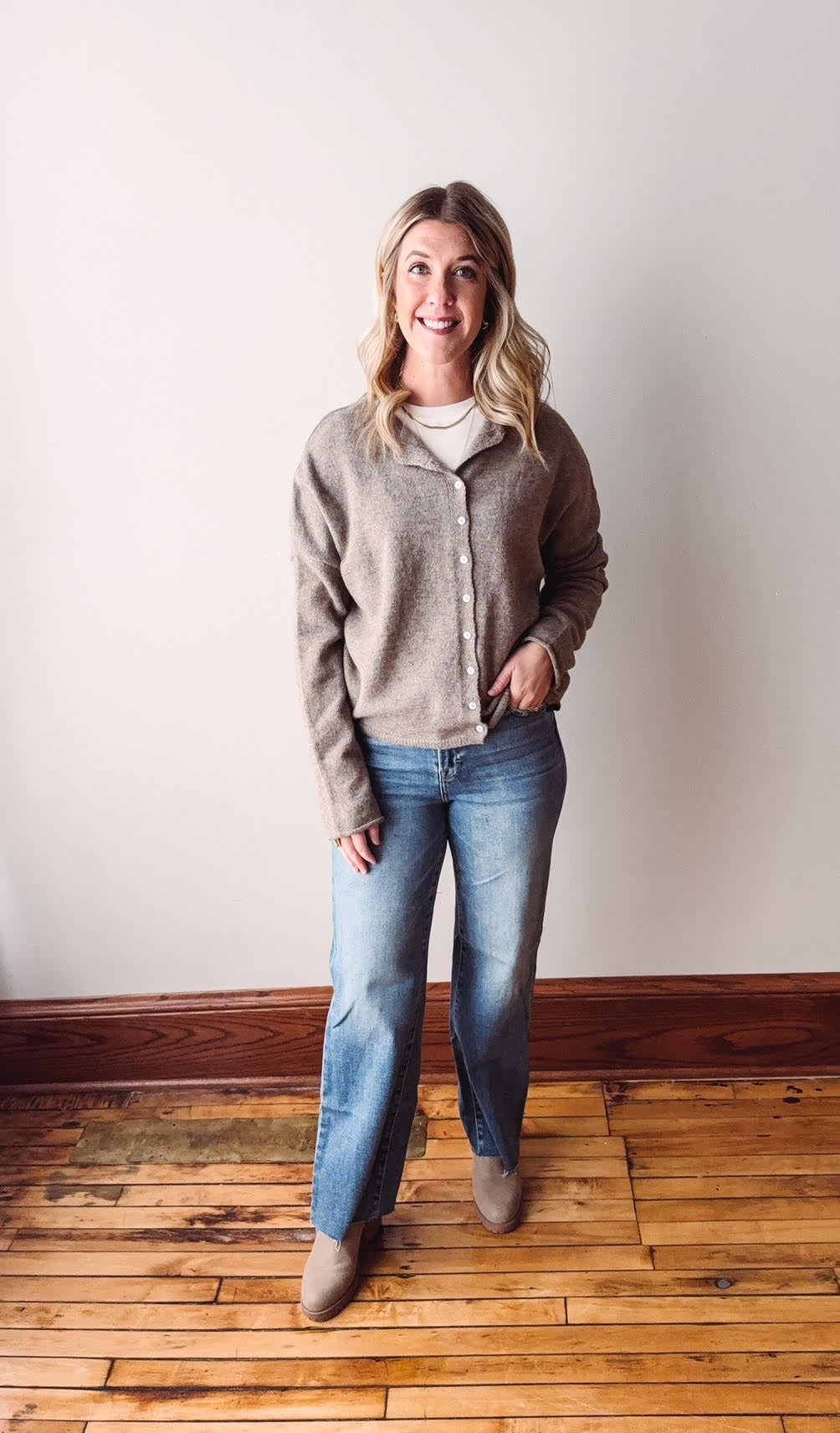 Woman wearing a gray sweater and blue jeans standing on a wooden floor with a white wall background