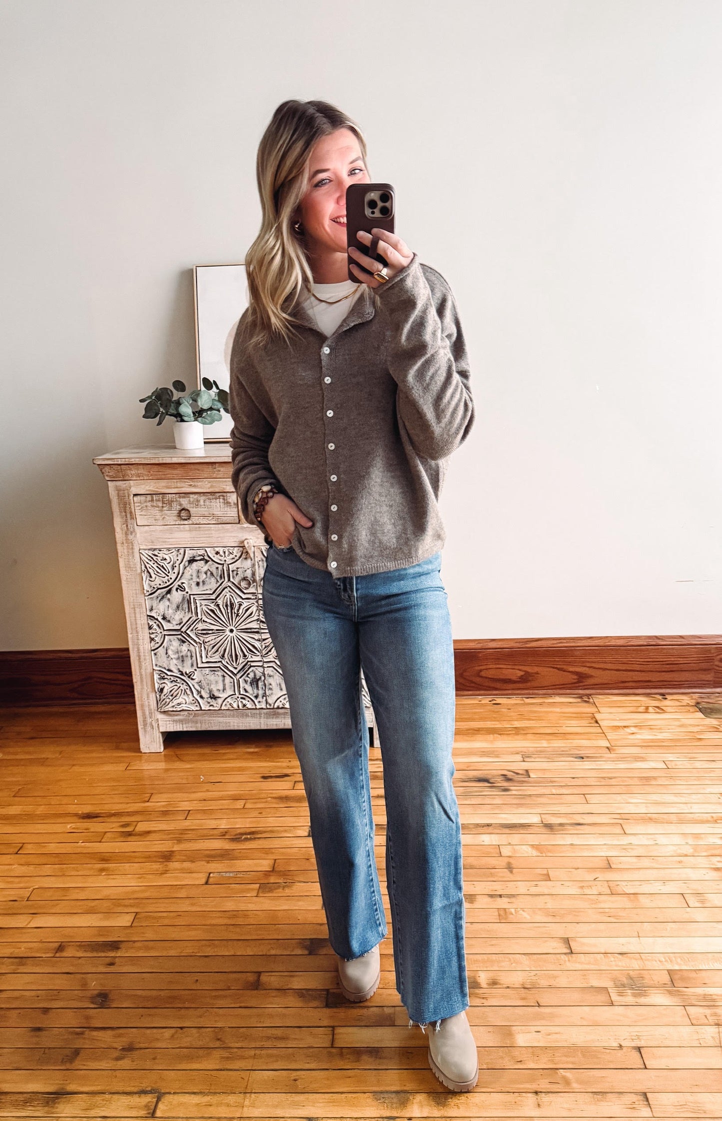 Woman taking a mirror selfie wearing a gray cardigan and blue jeans in a room with wooden flooring.