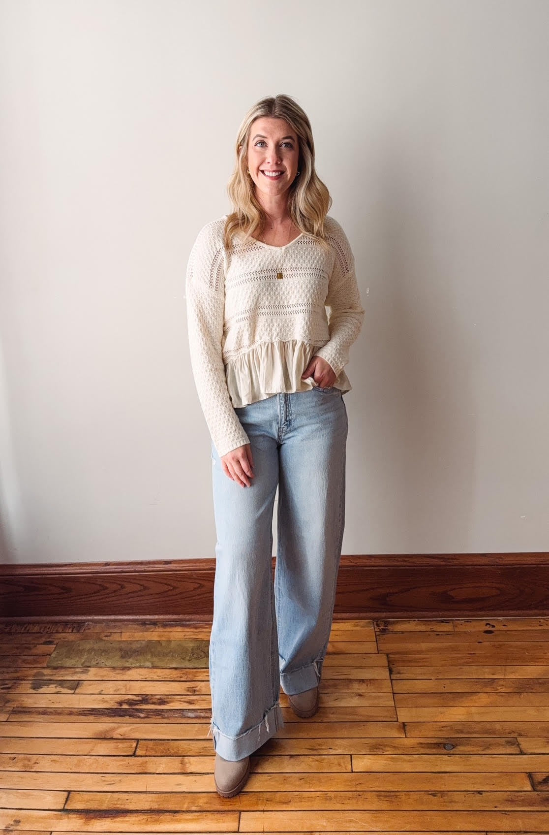 Woman wearing a beige sweater and light blue jeans standing on a wooden floor.