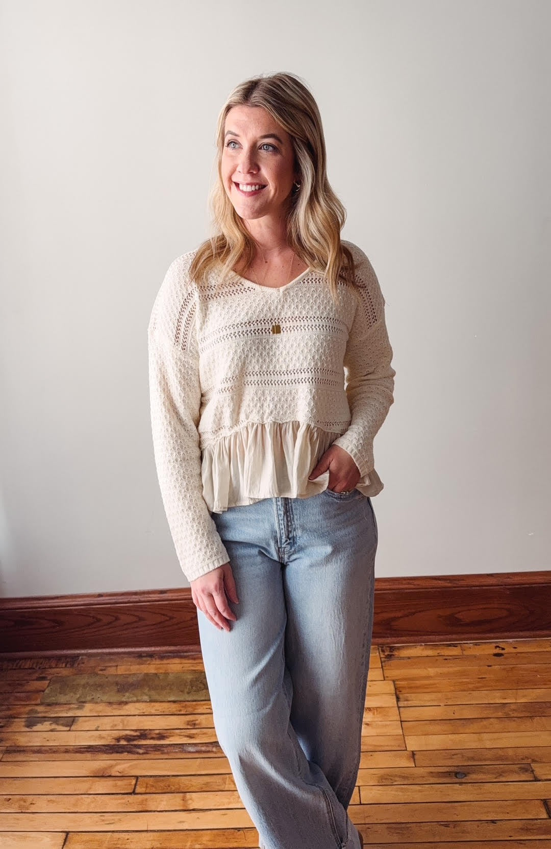Woman wearing a white lace top and light blue jeans standing on a wooden floor.