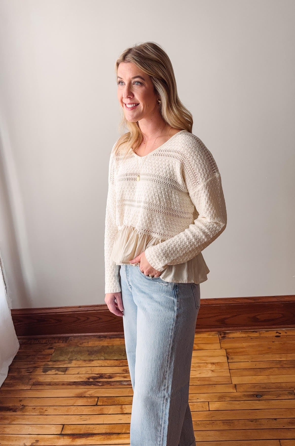 Woman wearing a cream sweater and light blue jeans standing in a room with wooden flooring and a white wall.