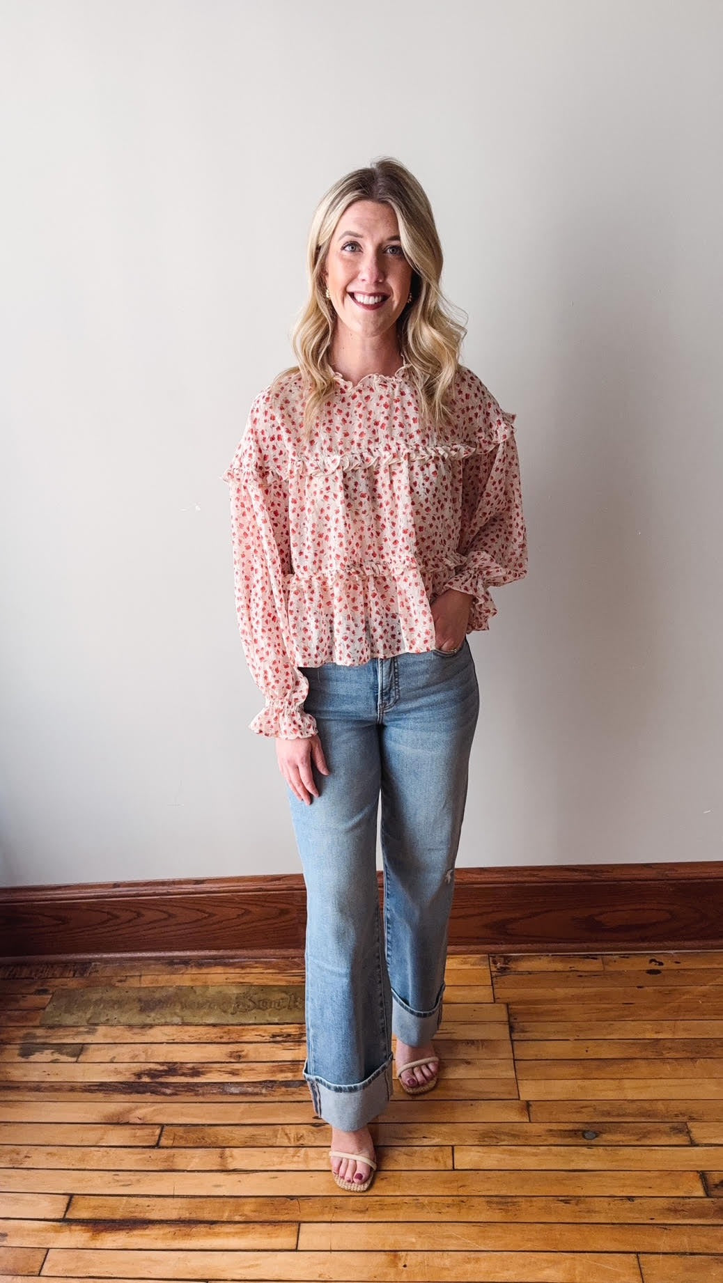 Woman wearing a pink floral blouse and blue jeans standing on a wooden floor.
