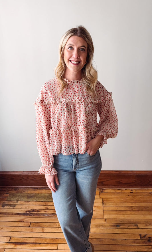 Woman wearing a pink floral blouse and blue jeans standing on a wooden floor with a white wall background.