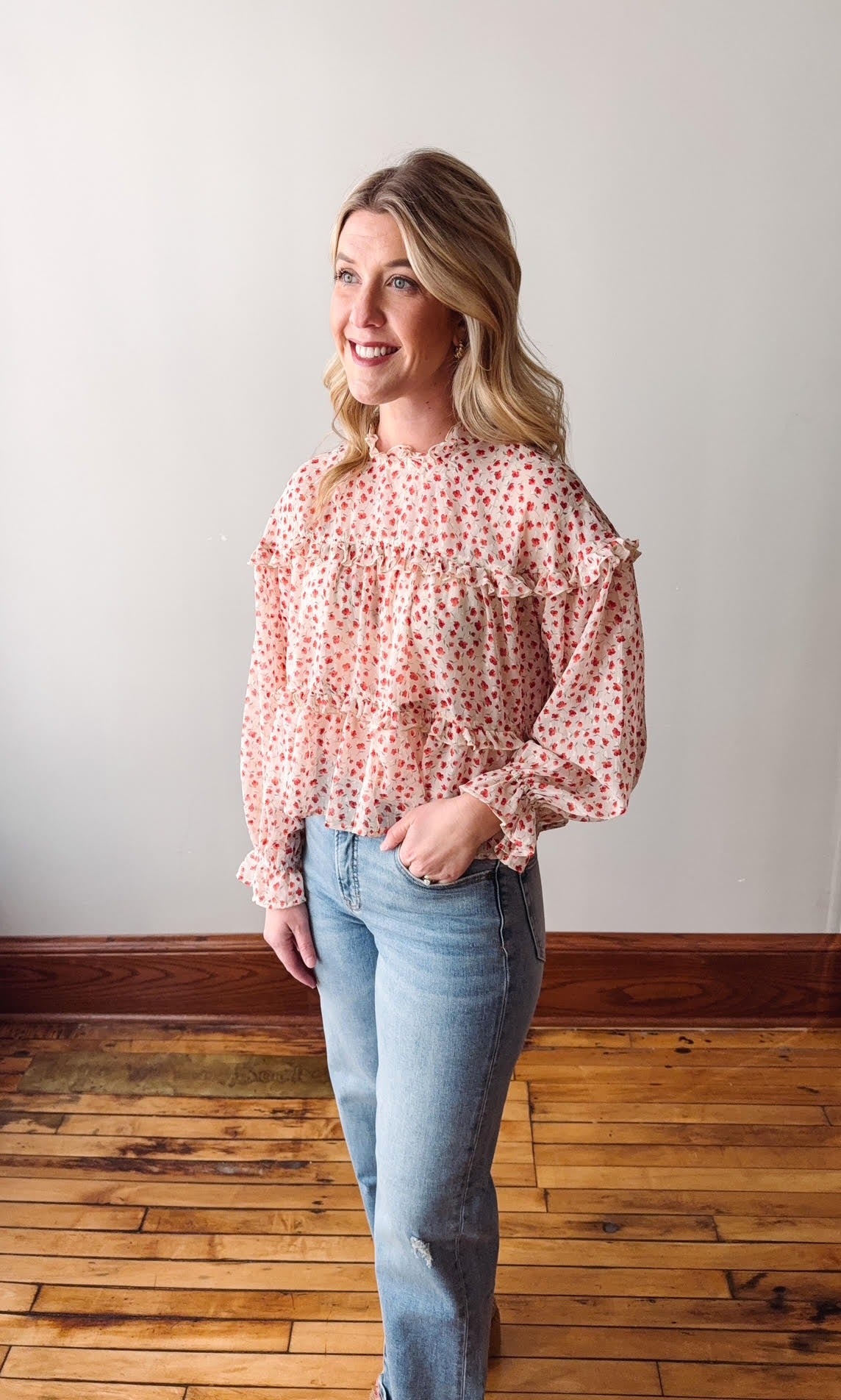 Woman wearing a pink floral blouse and blue jeans standing on a wooden floor.