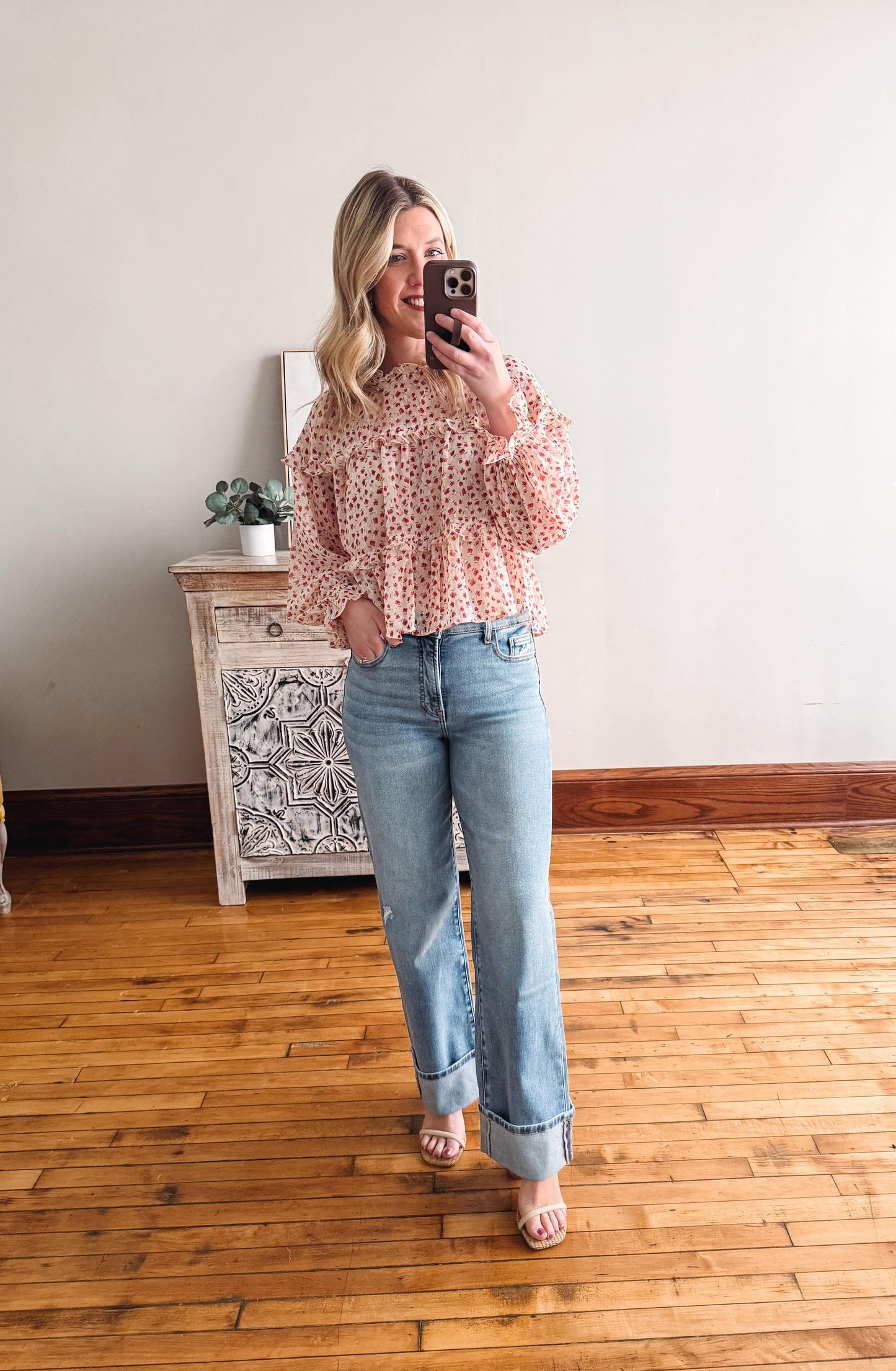 Woman taking a mirror selfie wearing a pink floral blouse and blue jeans in a room with wooden flooring.