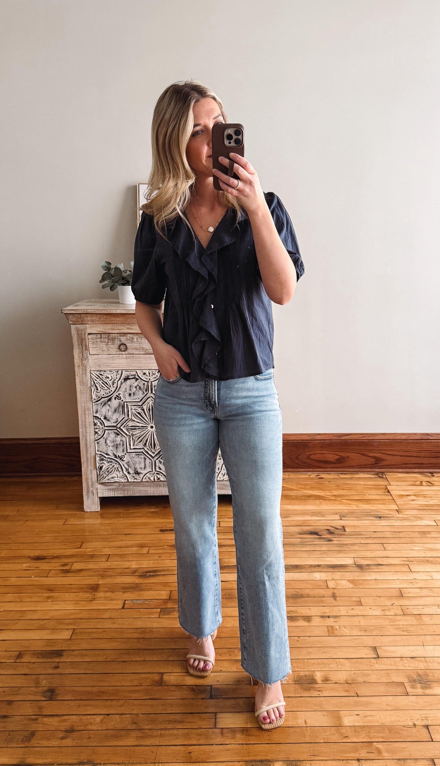 Woman taking a mirror selfie wearing a black top and light blue jeans in a room with wooden flooring.