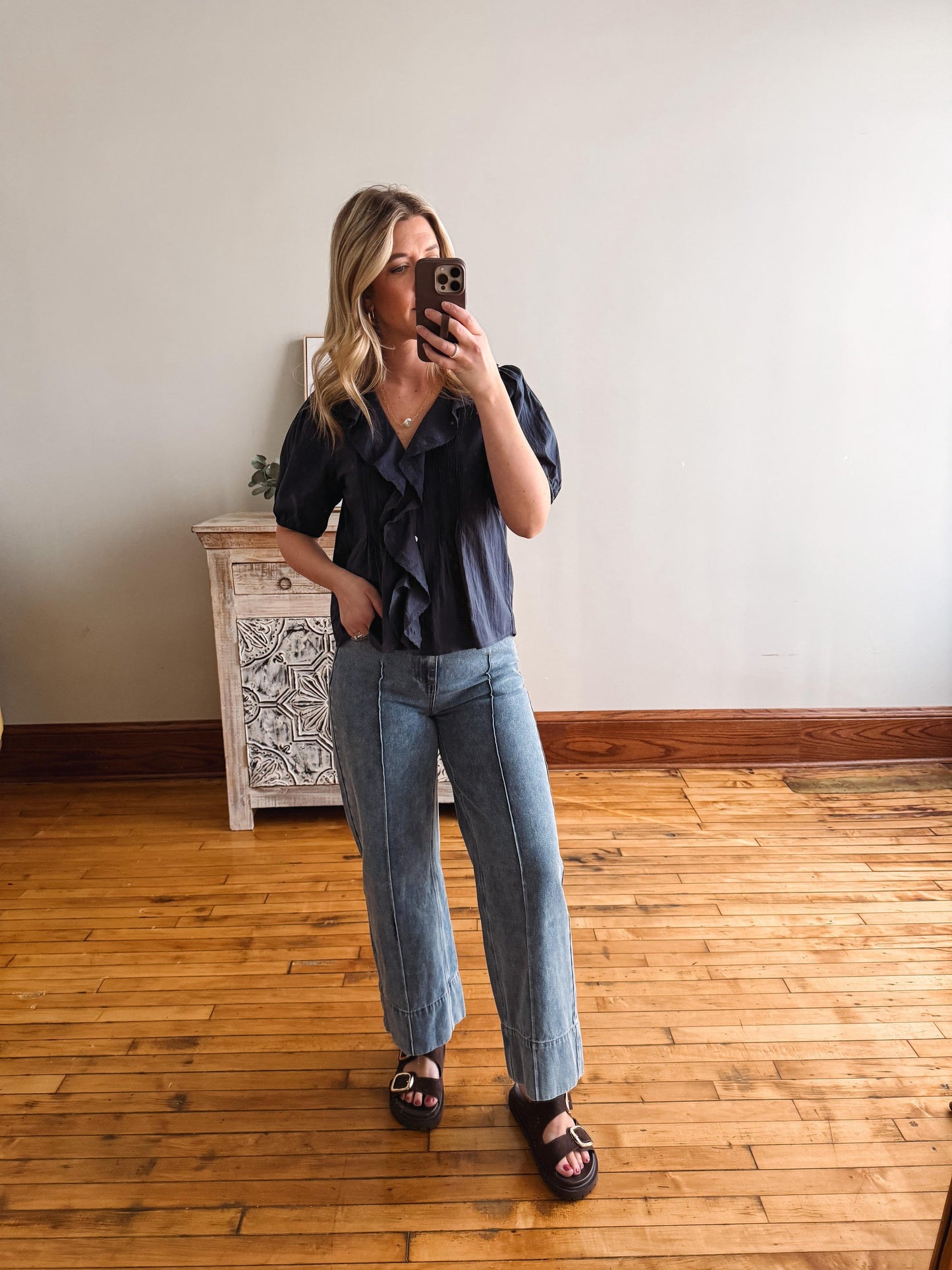 Woman taking a mirror selfie wearing a black shirt and blue jeans in a room with wooden flooring.
