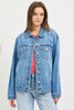 A woman wearing a light blue denim jacket over a coral-pink top and light-wash jeans, photographed against a plain light gray studio background.