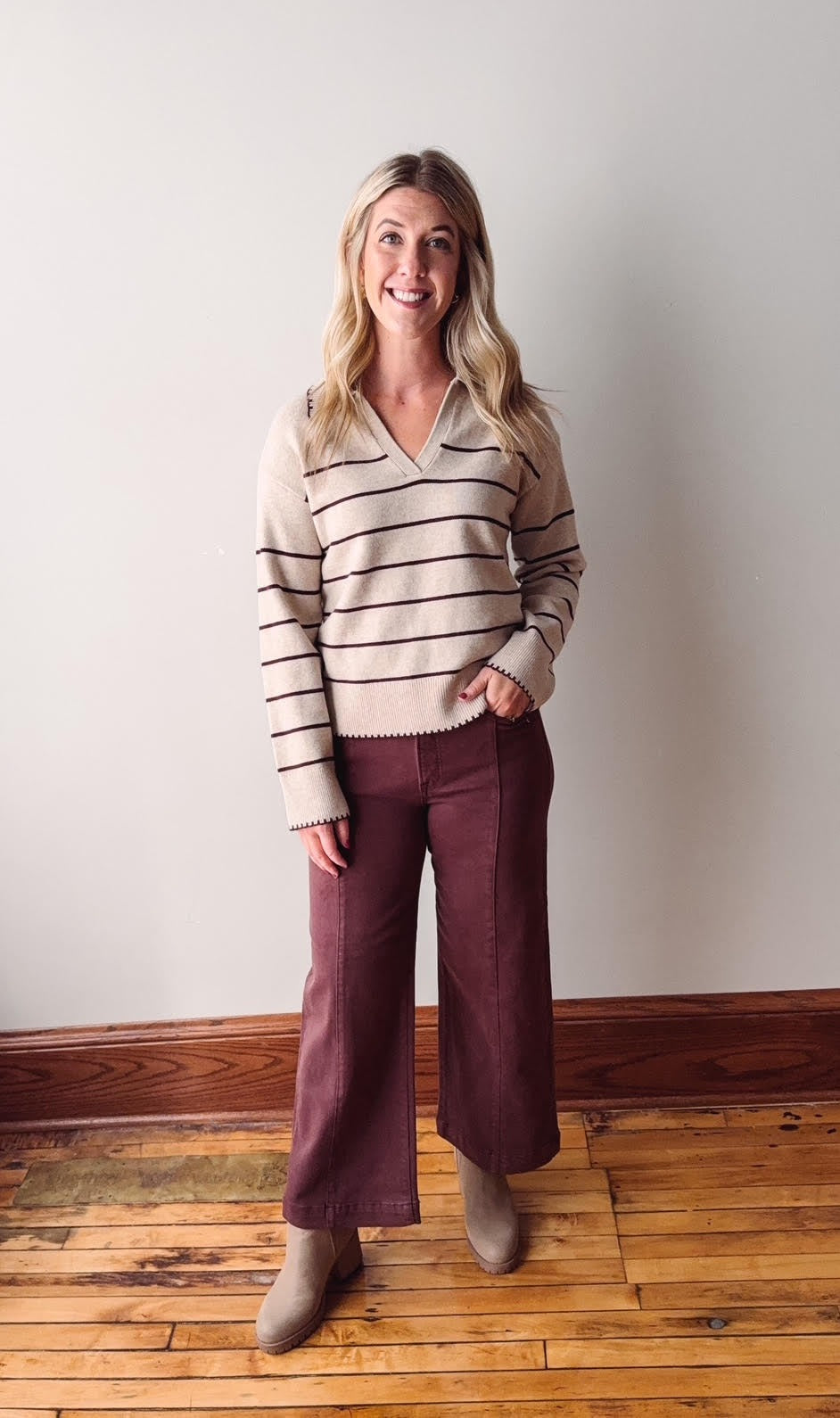 Woman wearing a striped sweater and burgundy pants standing against a plain wall.