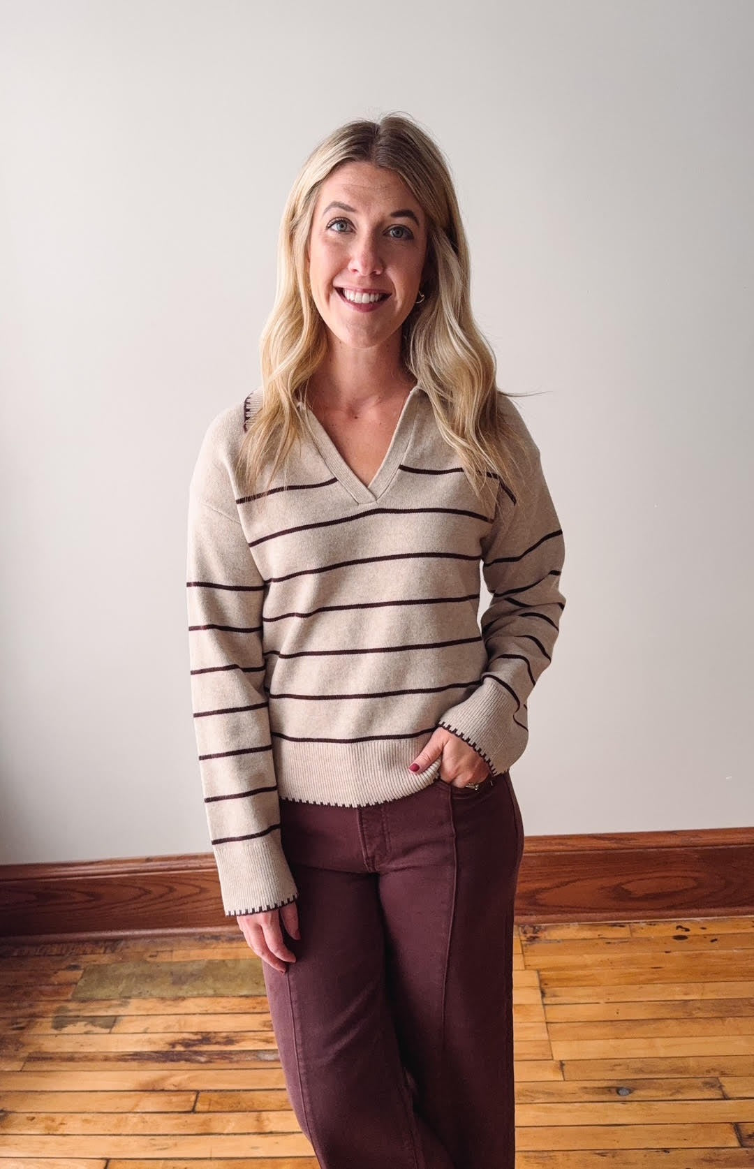 Woman wearing a striped sweater and burgundy pants standing against a plain wall.