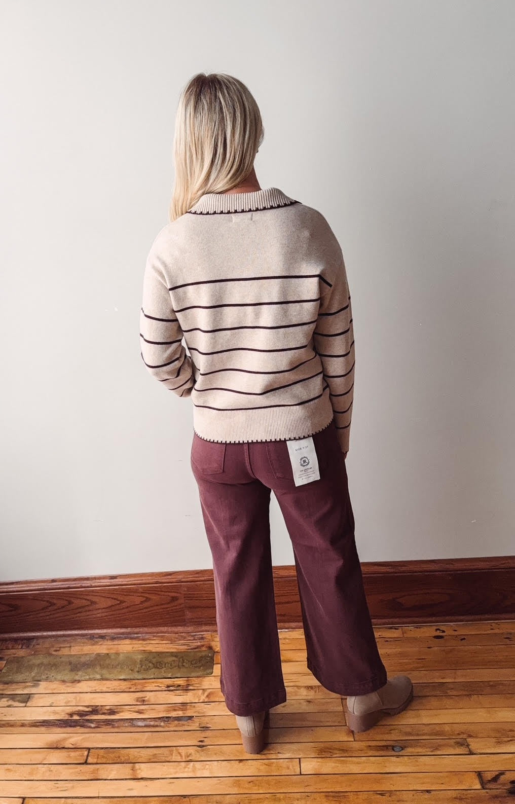Person wearing a striped sweater and burgundy pants standing against a plain wall.