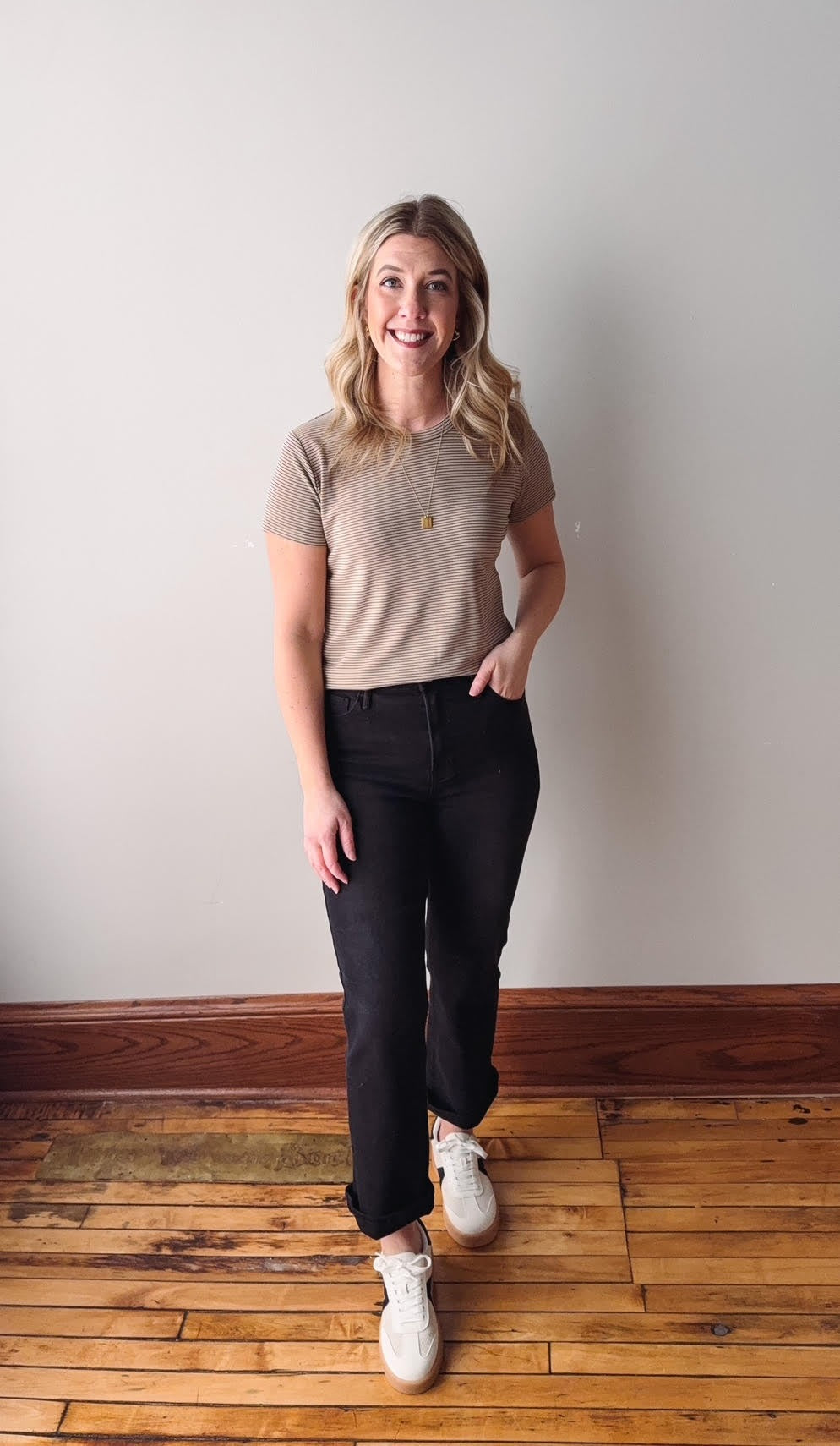 Woman wearing a beige t-shirt and black pants standing on a wooden floor with a white wall background