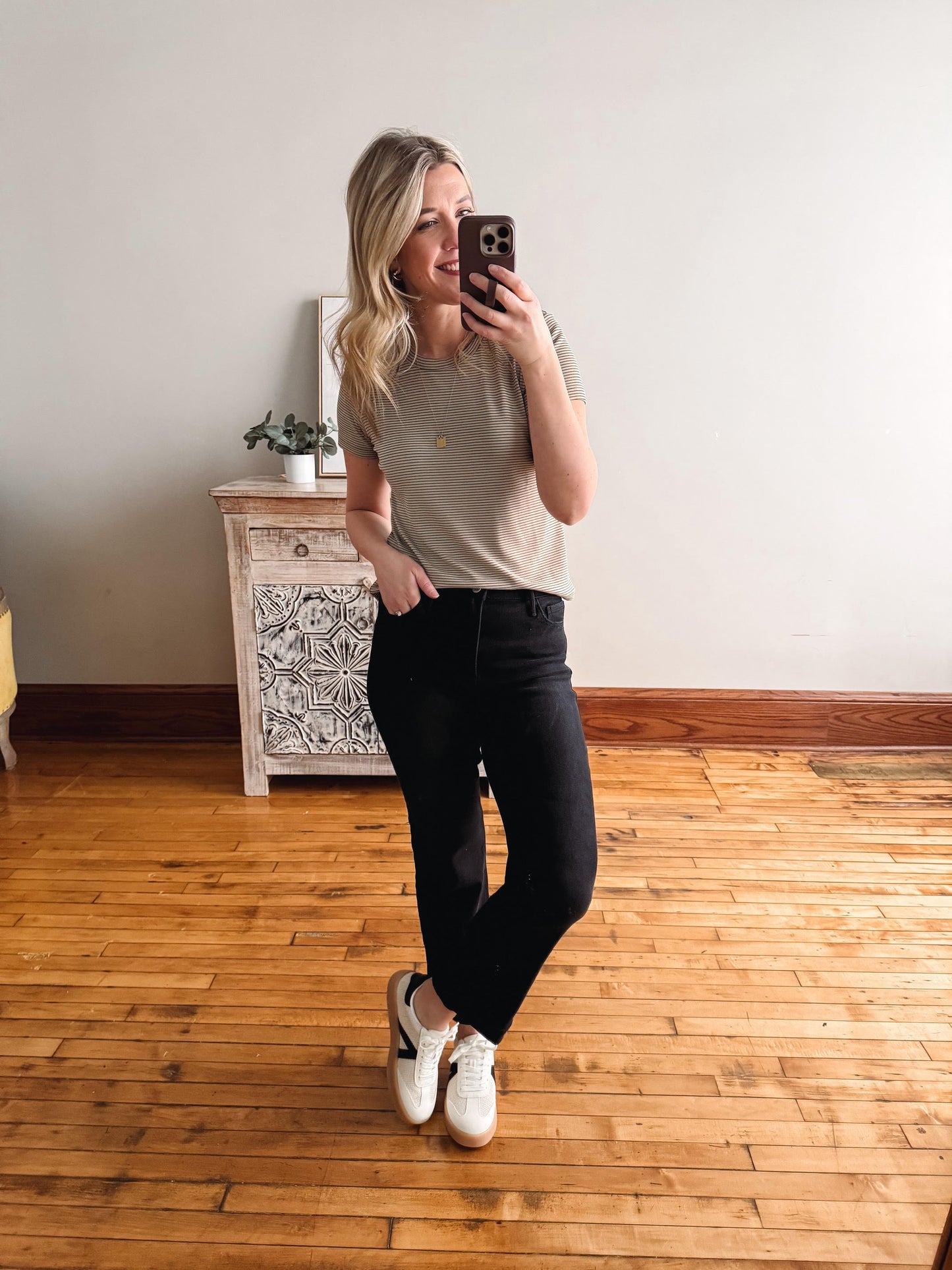 Woman taking a mirror selfie wearing a beige t-shirt and black pants in a room with wooden flooring.
